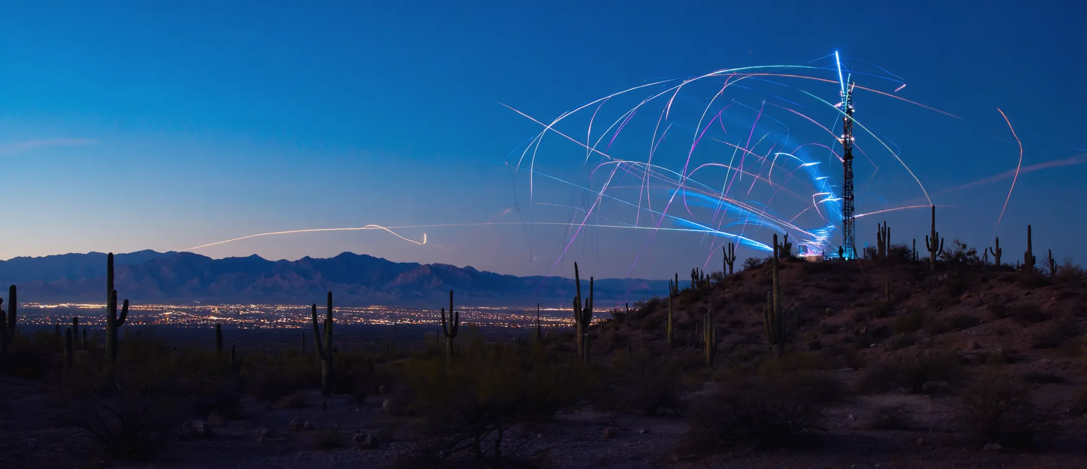 Phoenix skyline at dusk with streams of light arcing from a telecom tower over the Sonoran desert